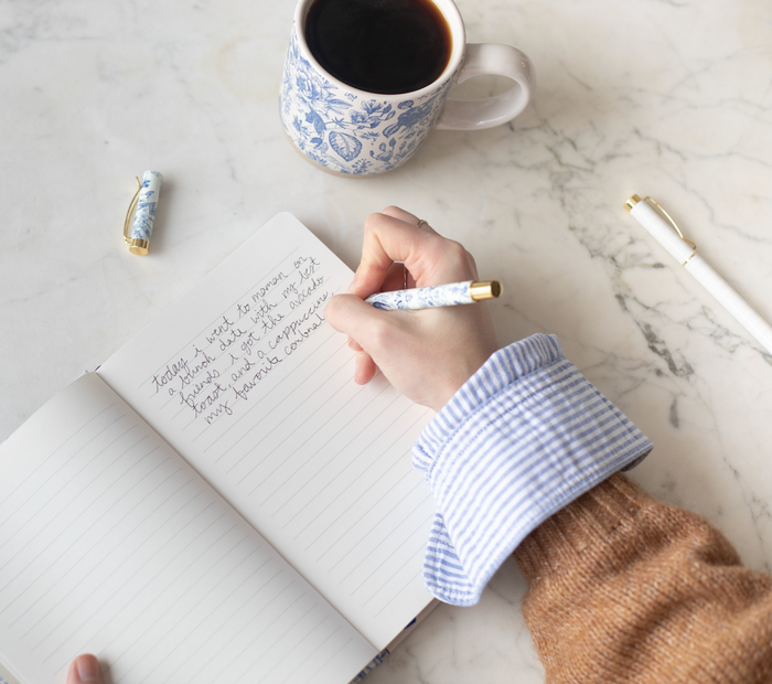 Person writing in a blue and white floral notebook with a matching blue and white floral pen, next to a blue and white floral stoneware mug.