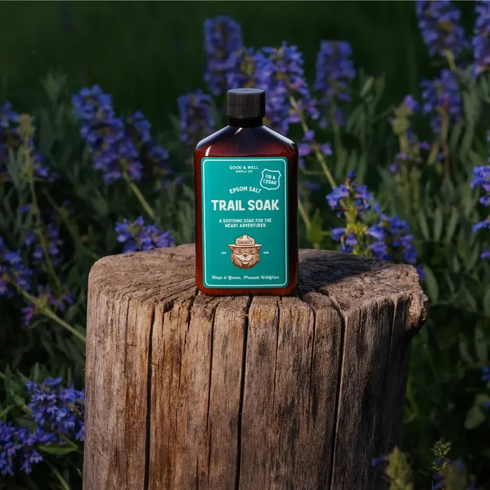 Good & Well Supply Co Smokey Bear Trail Soak Fir & Cedar bottle on tree stump, surrounded by lush grass and wildflowers outdoors.