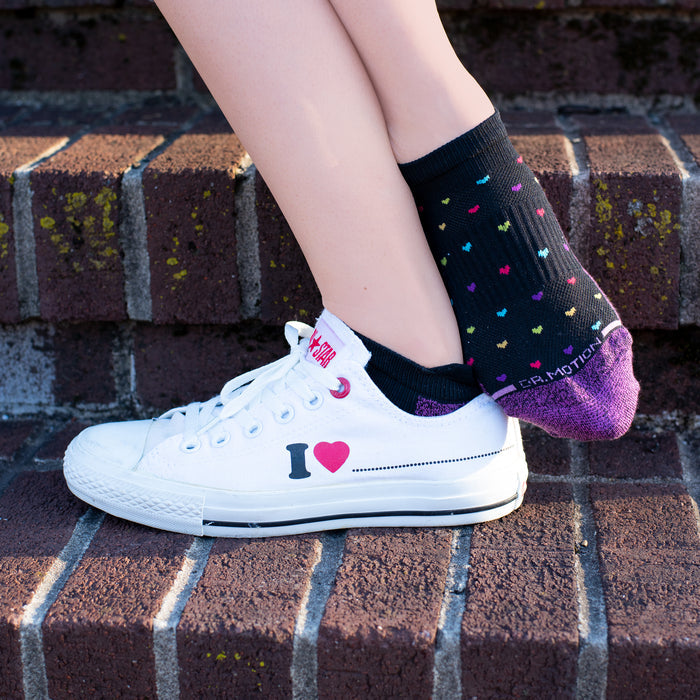 Ankle compression sock with colorful heart patterns worn with a white sneaker on a brick surface.