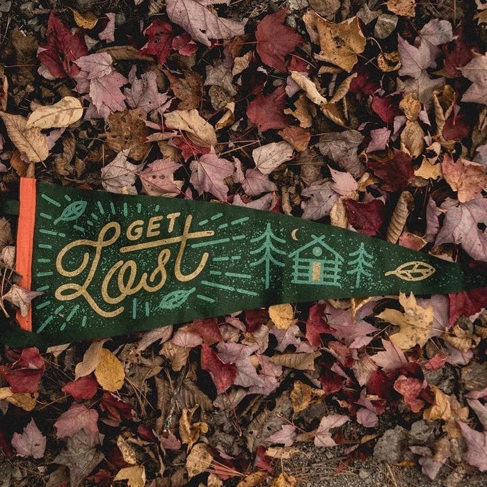 The Oxford Pennant Get Lost wool felt pennant, featuring a cabin graphic, lies on fallen autumn leaves in various shades of red, orange, and brown, inviting an outdoor adventure.