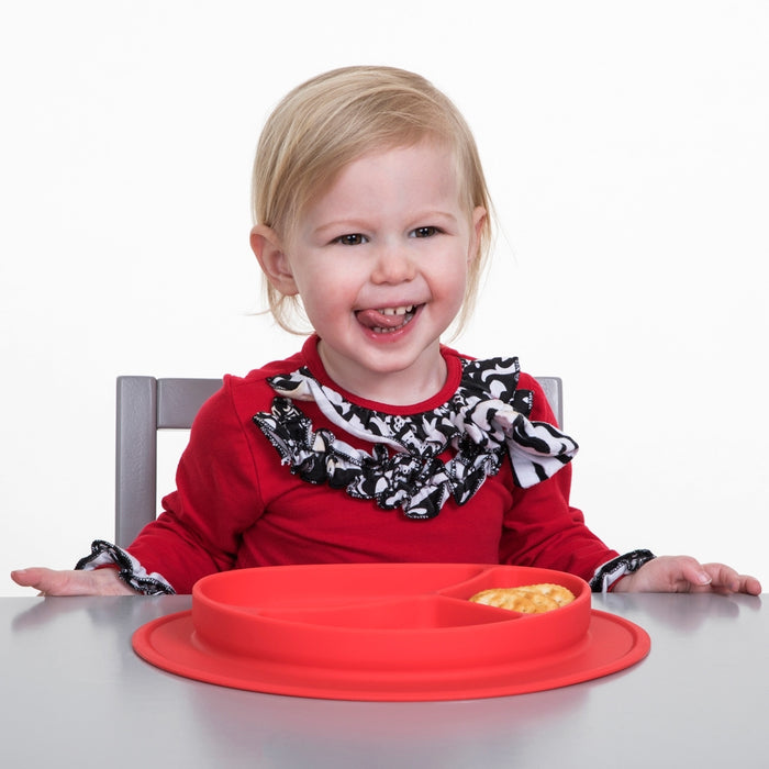A young child with blonde hair, wearing a red shirt with black and white ruffles, smiles at a table with a Sure Grip Miracle Mat 3-Section Plate in front of her, highlighting this playful silicone baby tableware filled with food.