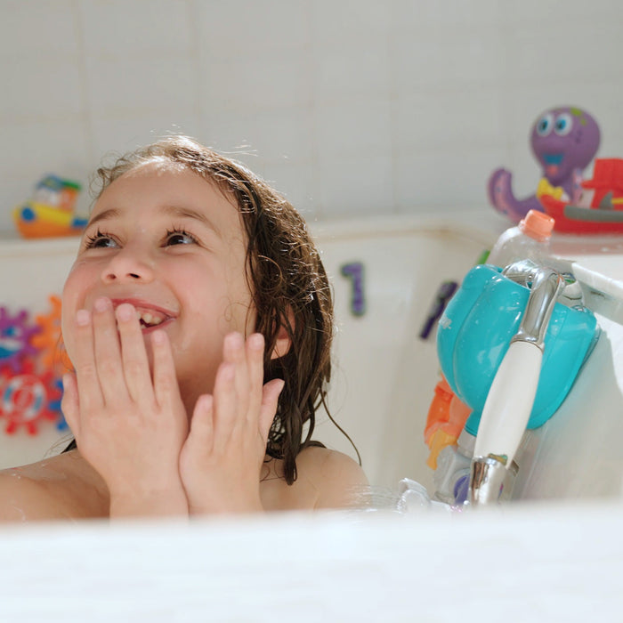 A young child with wet hair smiles in the tub, playing with Nuby’s Wacky Waterworks bath toy, which adds colorful fun and supports cognitive development during water play.