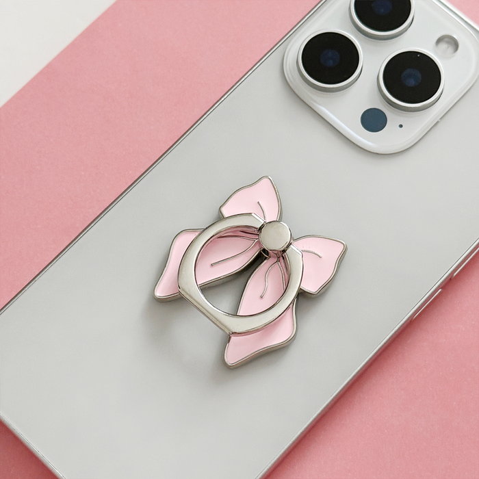 Pink bow Coconut Lane adhesive phone ring attached to a silver smartphone on a pink and white background.
