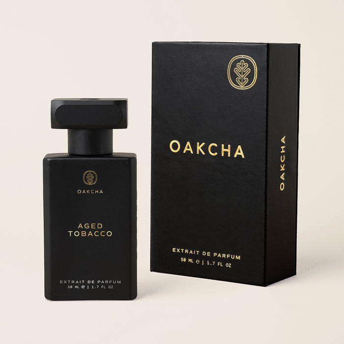 Front view of Aged Tobacco perfume bottle and box showcasing matte black bottle with gold logo beside matching black box on beige background