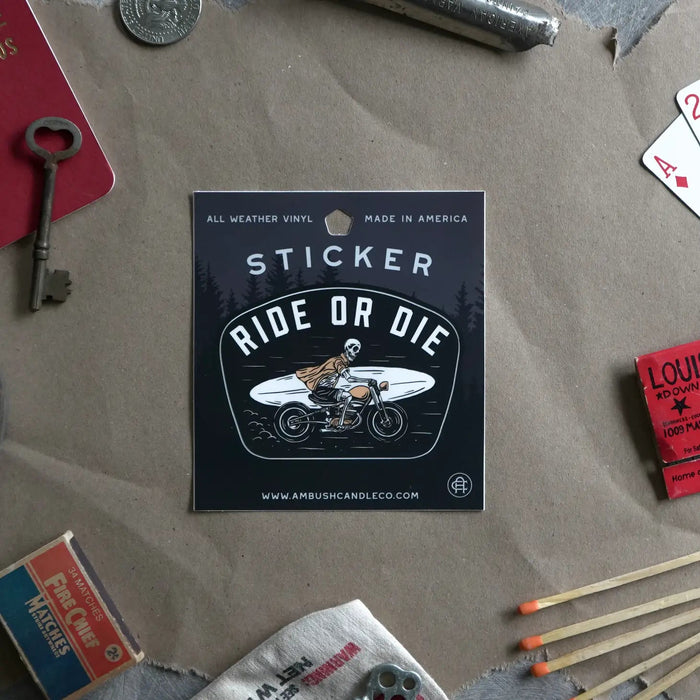 On a table, a weatherproof vinyl Ambush Candle Co "Ride or Die" sticker displays an image of a person on a bike, surrounded by various vintage items. This proudly made in the USA sticker adds character to the scene.