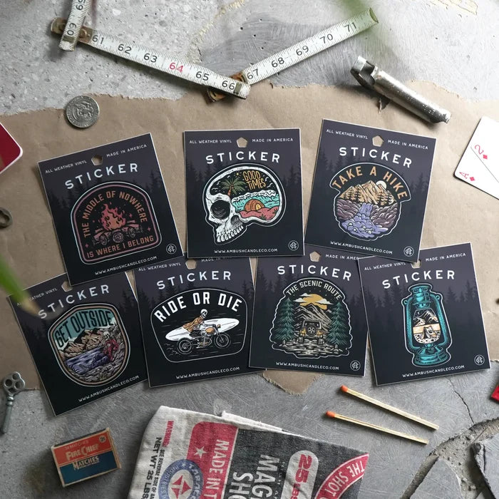 A weatherproof vinyl sticker from Ambush Candle Co, featuring the text "The Middle of No-Where is Where I Belong" and a campfire design, placed on a bed of fallen leaves.