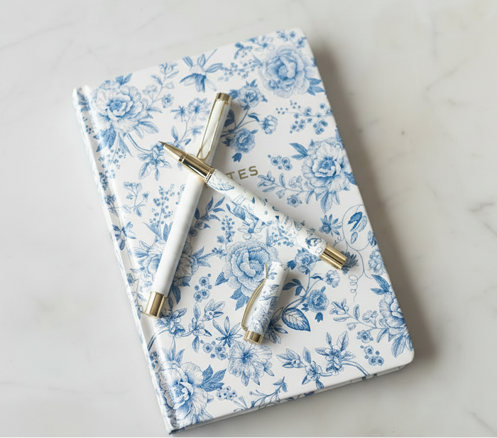 Matching set of a blue and white floral notebook, a blue and white floral pen, and a pink and white striped pen.