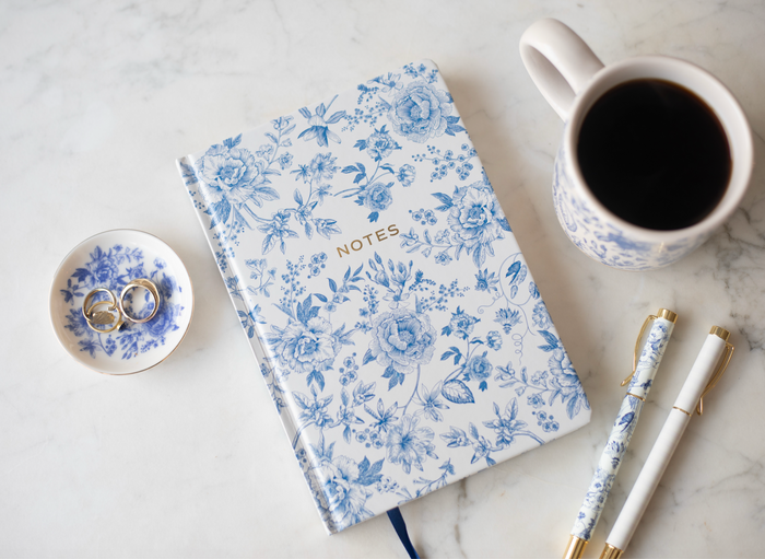 Matching set of a blue and white floral notebook, a blue and white floral pen, and a pink and white striped pen, next to the small blue and white floral jewelry dish.