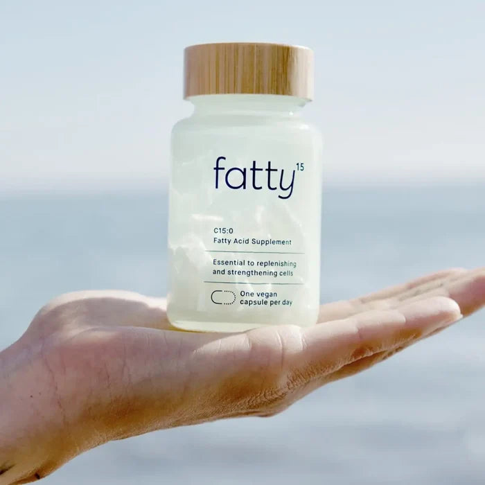 A hand holding a frosted supplement bottle labeled 'fatty15' with a bamboo-style lid, set against a soft blurred outdoor background.
