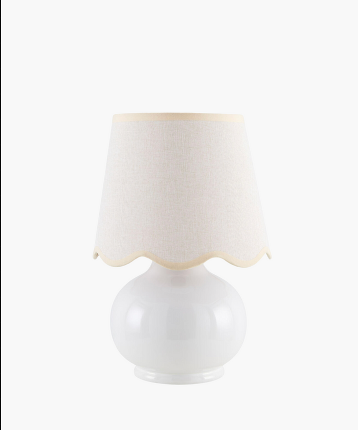 A white ceramic table lamp with a rounded base and a wavy-edged, light beige fabric shade. The design is simple, elegant, and modern.