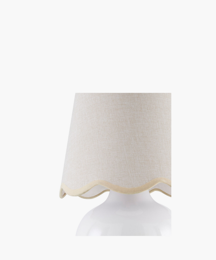 A white ceramic table lamp with a rounded base and a wavy-edged, light beige fabric shade. The design is simple, elegant, and modern.