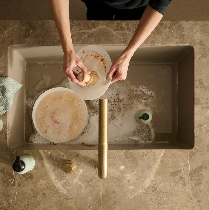 Guests on Earth Monogram Dish Brush scrubs a stained plate over a soapy sink in a warm, modern kitchen