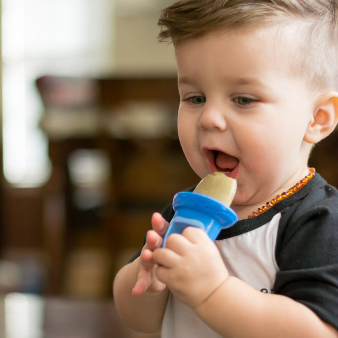 A young child with short light brown hair enjoys a frozen treat made with the Fruitsicles Ice Pop Tray, using a blue-handled pop indoors. The child wears a black and white shirt and beaded necklace against a blurred background.