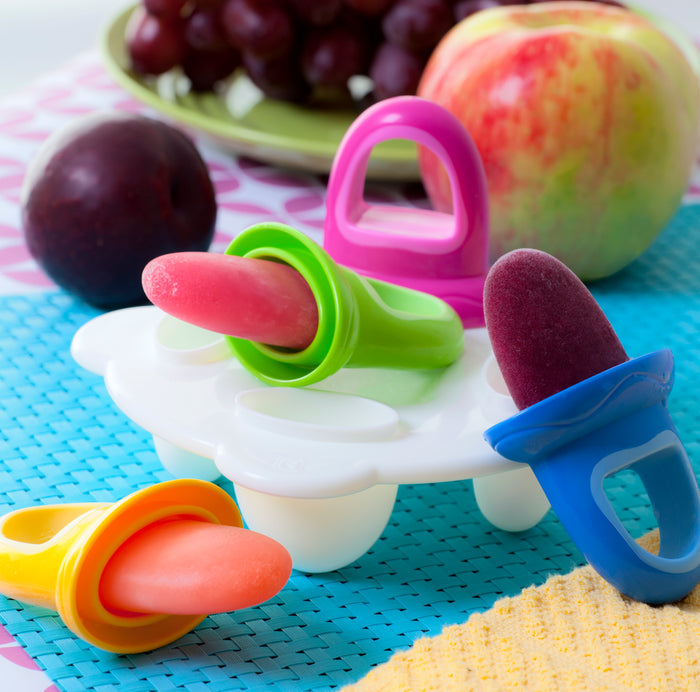 Homemade frozen fruit pops in ring-shaped molds from the Fruitsicles Ice Pop Tray rest on a white holder, with grapes, plum, and apple behind—ideal for teething babies and vibrant fun on blue and pink backgrounds.