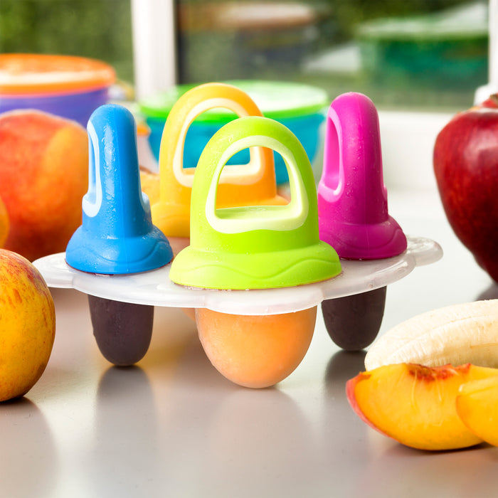 Four vibrant frozen pops in the Fruitsicles Ice Pop Tray rest on a table with peaches, apples, bananas, and nectarines nearby—perfect for teething babies. A window and greenery are in the background.