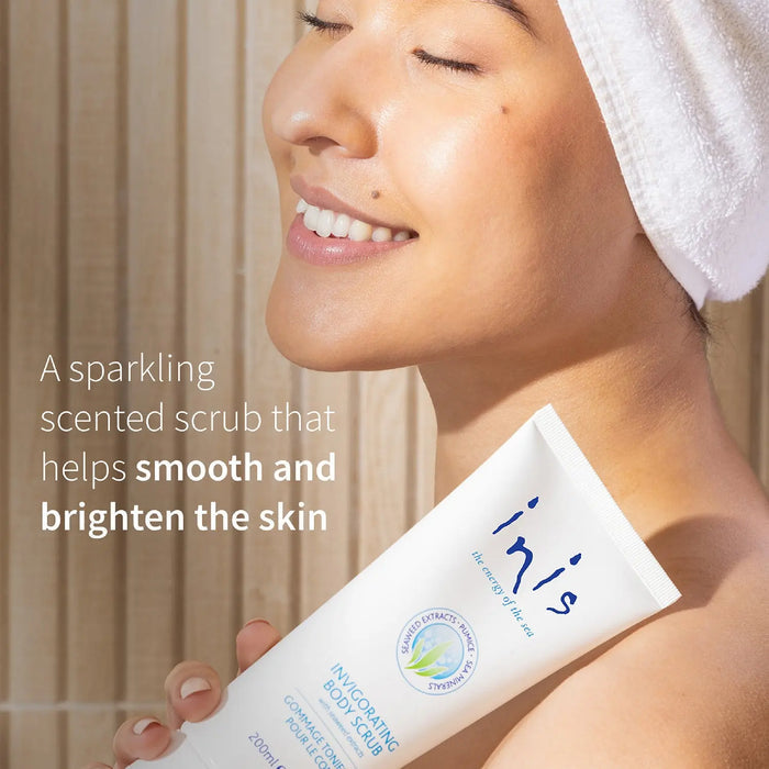 A smiling woman with a white towel wrapped around her hair holds an Inis Invigorating Body Scrub tube. Text on the left reads, 'A sparkling scented scrub that helps smooth and brighten the skin.