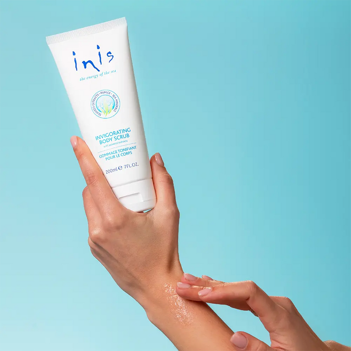 Hands hold Inis Invigorating Body Scrub tube against a light blue background, while another hand applies the white cream to an arm.