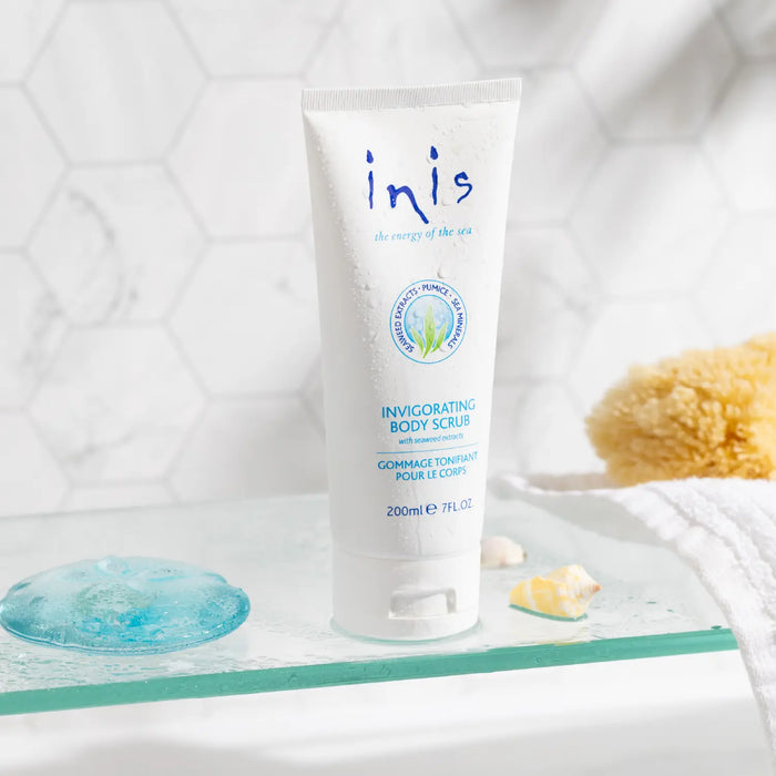 A 200ml white tube of inis Invigorating Body Scrub stands on a glass tray in a bright bathroom setting with a yellow sponge, and a white towel.
