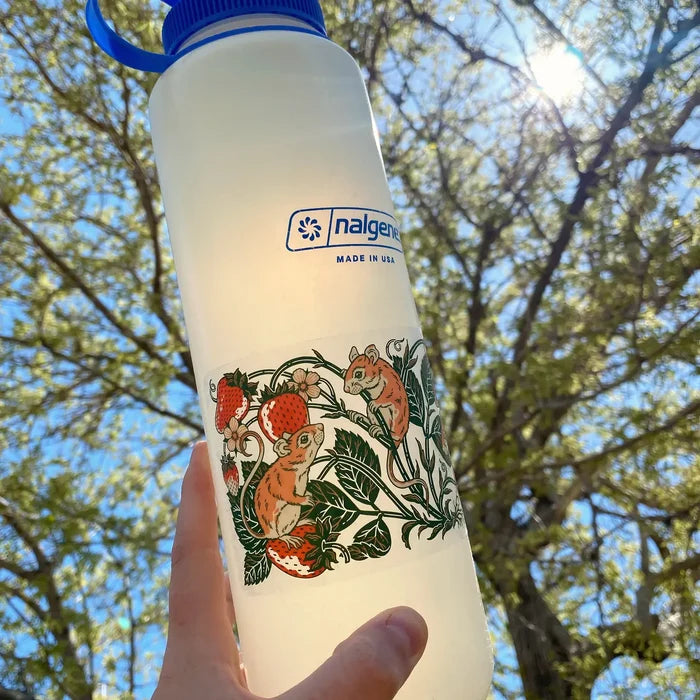 Hand holding Nalgene bottle with Kate O’Hara Mice and Strawberries sticker; trees, nature, sunlit branches in background.
