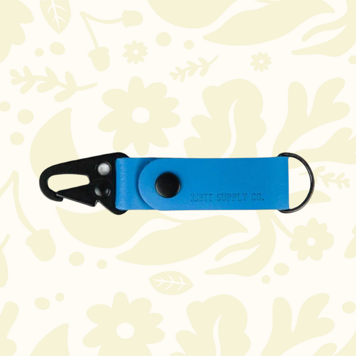 Blue Kjøtt Forager Provisions Leather Key Fob with black clip and ring, shown on a beige floral background.