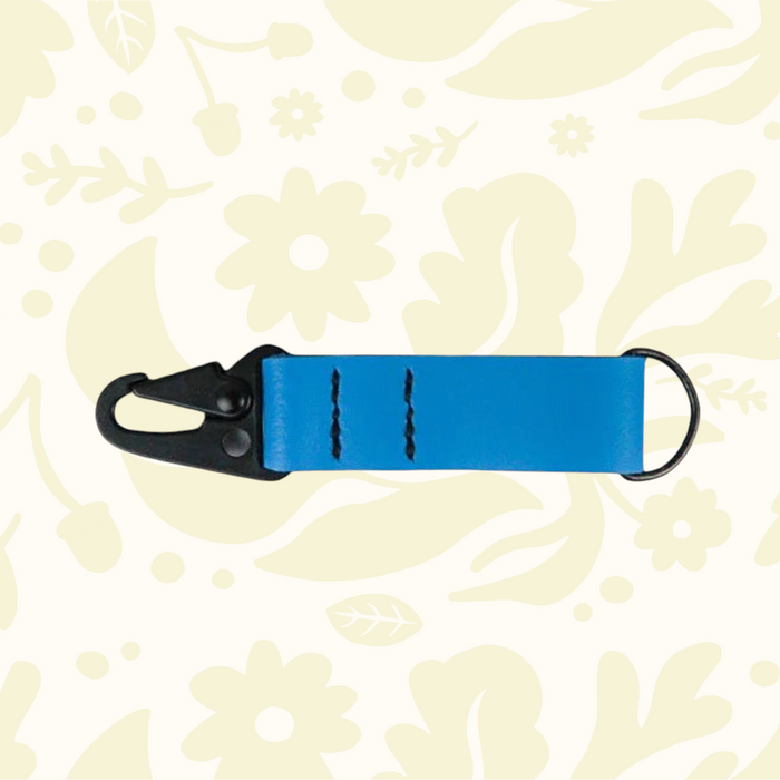 Kjøtt Forager Provisions blue leather key fob with black metal clip, stylishly displayed on a beige floral background.