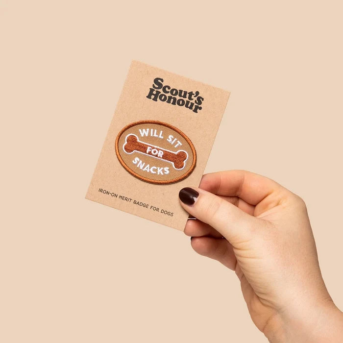 Hand holds Scout’s Honour Will Sit For Snacks Dog Patch, bone-shaped embroidered badge for dogs, obedience training.