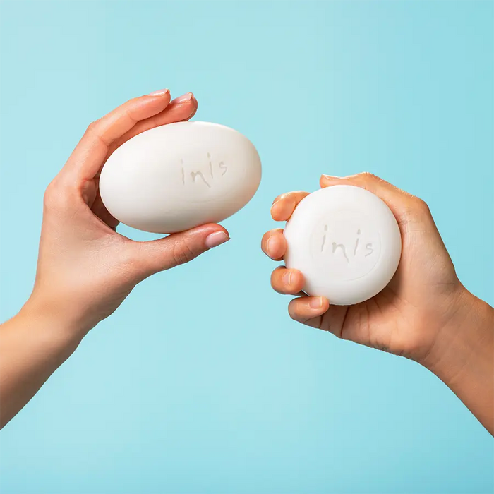 Two hands holding white Inis soap bars against a light blue background