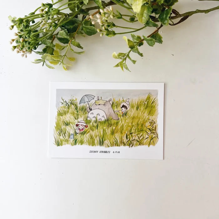 A Seenee Scribbles Art Print measuring 4 x 6 inches, featuring a charming watercolor illustration of Totoro and friends in a grassy field, reminiscent of classic Studio Ghibli scenes. Delicate flowers adorn the upper left corner, enhancing the whimsical beauty of this Totoro In The Fields print.
