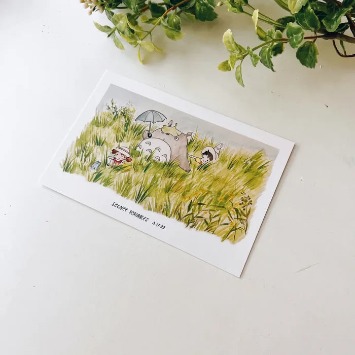 A Seenee Scribbles Art Print measuring 4 x 6 inches, featuring a charming watercolor illustration of Totoro and friends in a grassy field, reminiscent of classic Studio Ghibli scenes. Delicate flowers adorn the upper left corner, enhancing the whimsical beauty of this Totoro In The Fields print.