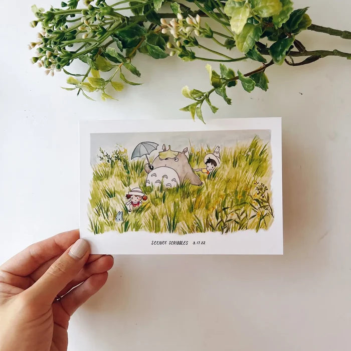 A Seenee Scribbles Art Print measuring 4 x 6 inches, featuring a charming watercolor illustration of Totoro and friends in a grassy field, reminiscent of classic Studio Ghibli scenes. Delicate flowers adorn the upper left corner, enhancing the whimsical beauty of this Totoro In The Fields print.