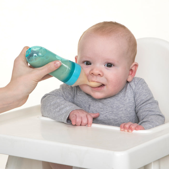 A baby in a high chair, dressed in a gray onesie, is being fed by an adult with the Nuby Silicone Squeeze Feeder with Cover. The baby looks ahead contentedly, the feeder's silicone spoon gently in their mouth.