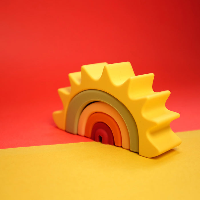 The Silicone Stacking Sun Toy features rainbow arches in yellow, orange, pink, and gray. Its spiked design resembles sun rays, enhancing cognitive development. Shown on a yellow surface with a red background.