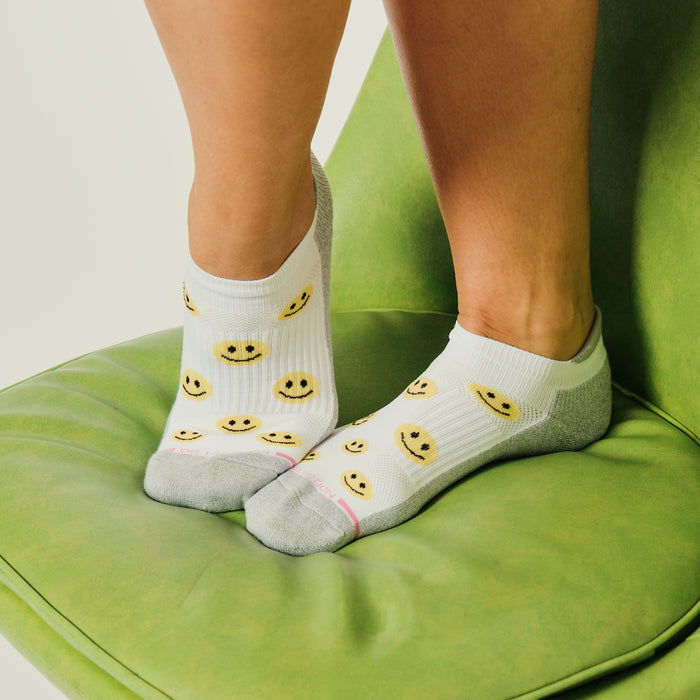 Ankle compression socks with a white base and yellow smiley face patterns, worn on feet resting on a green chair.
