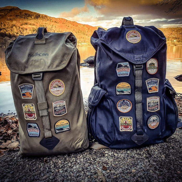 Two backpacks with Scafell Pike patches by Adventure Patch Company on Lake District shore at sunset, hiking and travel gear.
