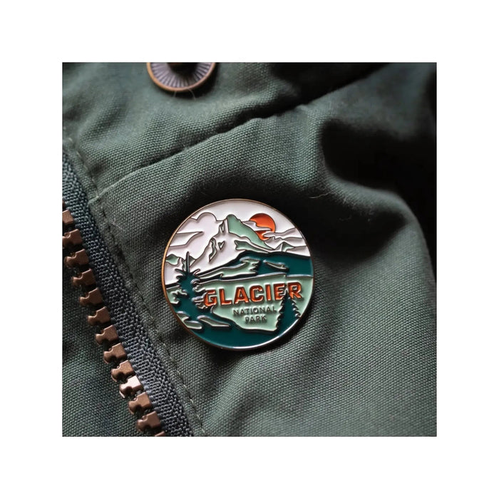 The Landmark Project Glacier National Park Pin is a round enamel pin featuring a mountain and river design with the text "GLACIER NATIONAL PARK." Attached to a green fabric backing, this pin celebrates Glacier National Park and highlights our dedication to sustainable packaging, making it an ideal addition for enamel pin collectors.