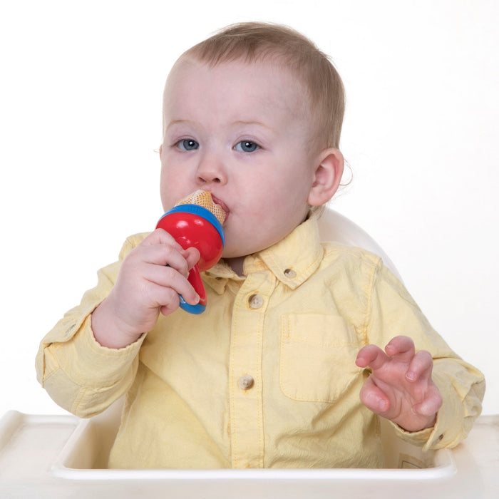 A baby in a yellow shirt sits in a high chair, holding and sucking on The Nibbler Mesh Net Feeder to try new foods. The background is plain white.