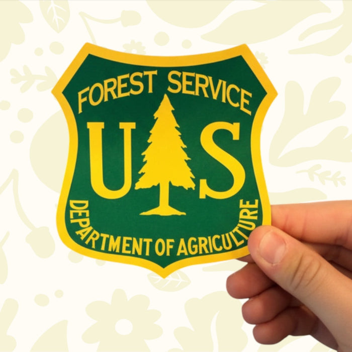 Hand holds The Sawmill Shop Made in USA US Forest Service Wood Shield 5x3” against a patterned backdrop, closeup.