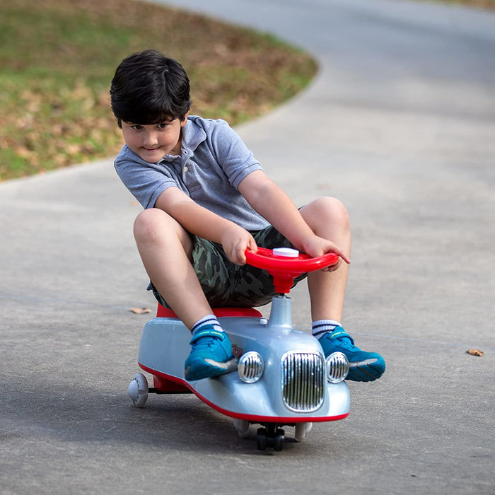 A boy with dark hair and a gray polo shirt rides a blue and red Twist N' Ride Classic Car with light-up wheels, gripping the steering wheel and leaning playfully as he cruises outdoors on a paved path.