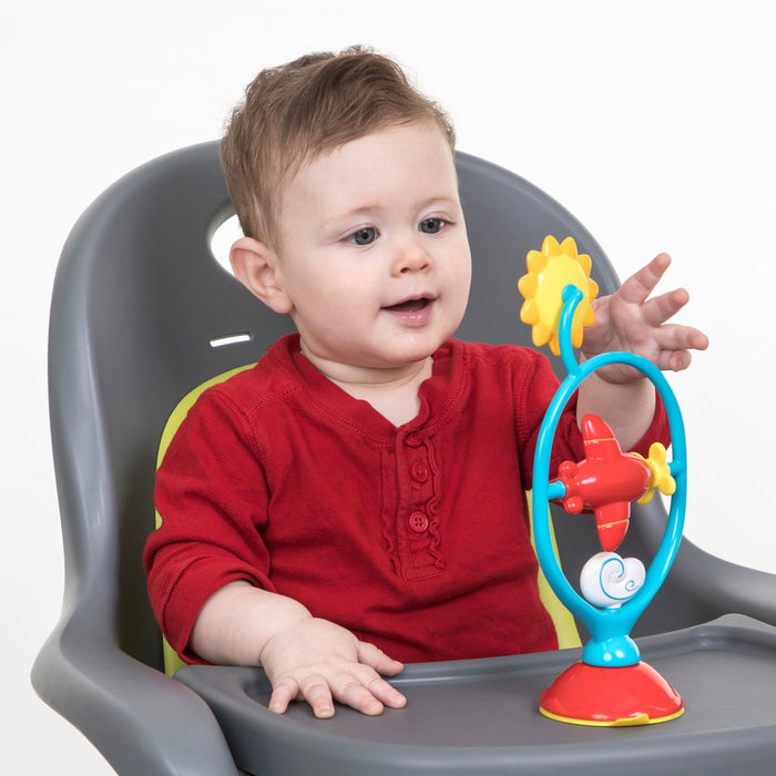 A baby in a red shirt sits in a high chair, playing with the Whirly Wings interactive toy, spinning the red airplane on its blue and yellow frame for sensory development.