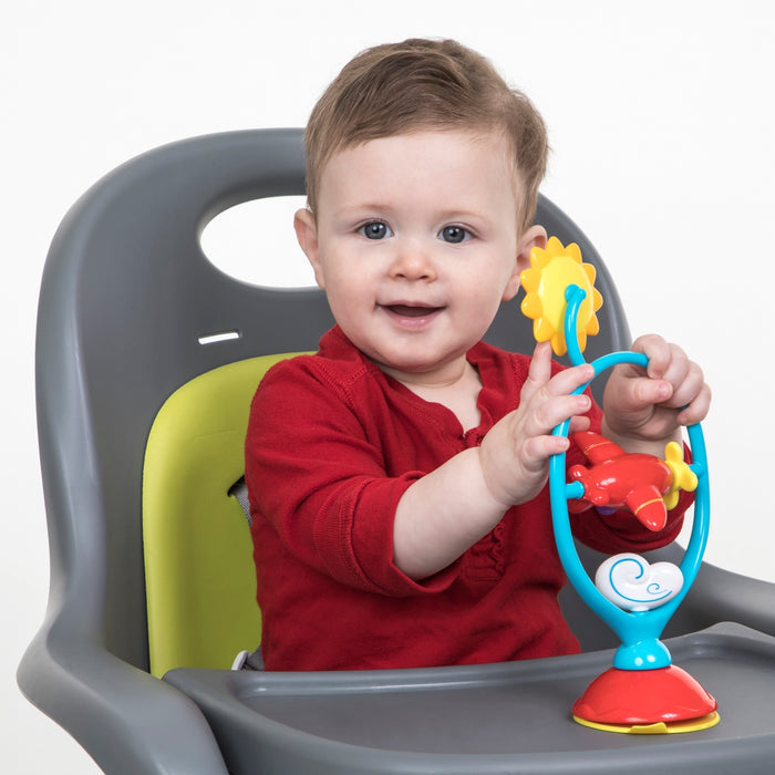 A smiling baby with short light brown hair, in a red shirt, sits in a high chair and plays with Whirly Wings, an interactive baby toy featuring a sturdy suction base that aids sensory development.
