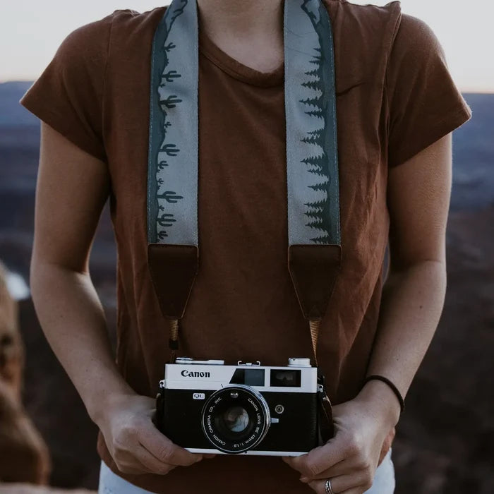Outdoor photographer in brown shirt holds Canon camera with Wildtree Landscape Strap, featuring cacti, trees, and mountains.