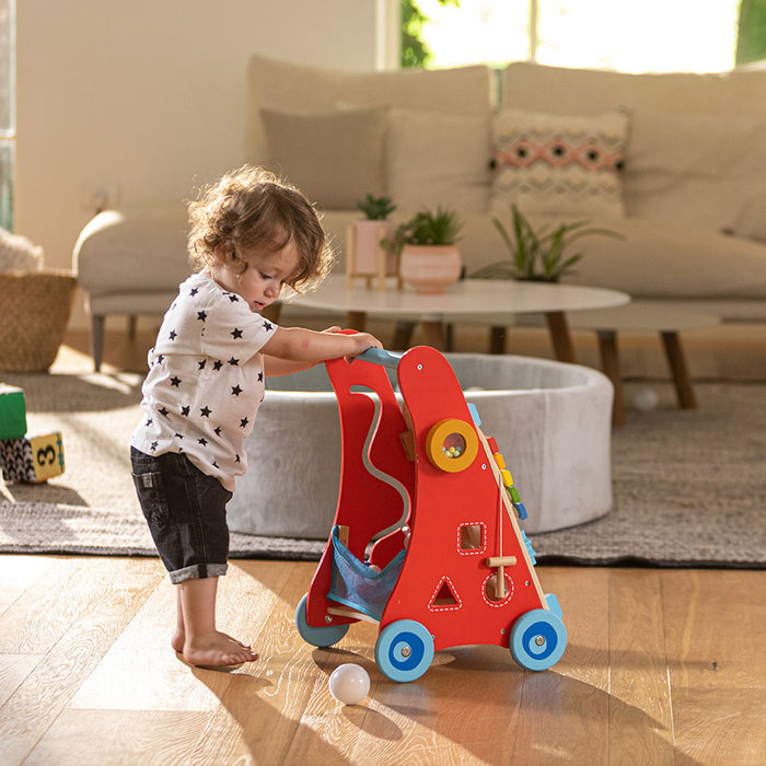 A toddler with curly hair plays with a colorful activity walker in a sunlit living room, standing on a wooden floor near a white rug and modern Montessori-inspired furniture.